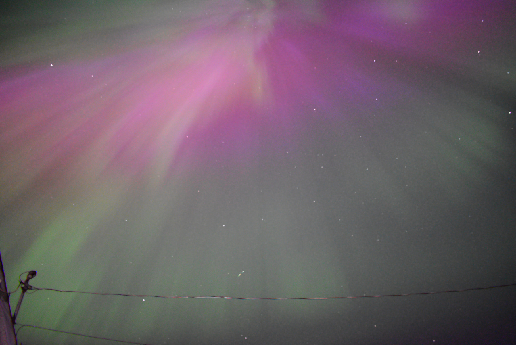 A long exposure photography of the Aurora as seen from British Columbia, Canada. A large number of bright pink and green lights are seen emanating from a radiant point high in the sky. Some bright stars can be seen in the background.